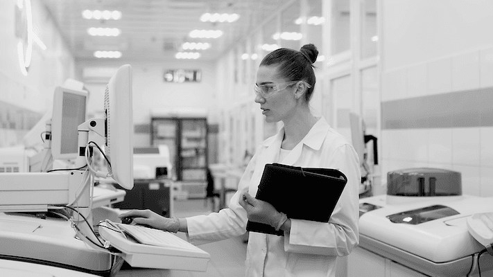 Woman in lab coat and goggles holding a binder looking at mobile medical computer